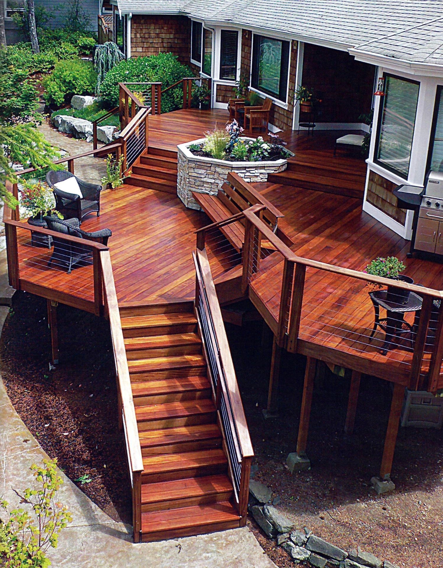 Wooden multi-level deck with stairs and seating, surrounded by lush greenery and garden plants.